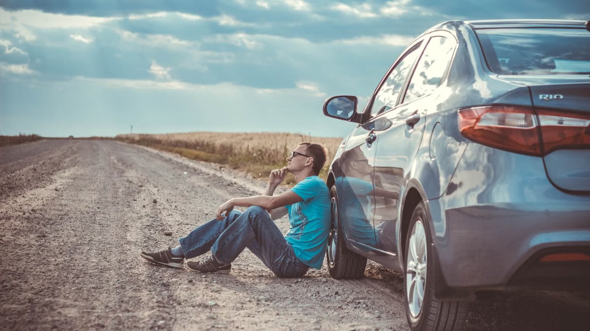 Man sitting by a car on a remote gravel road under a cloudy sky, deep in thought.